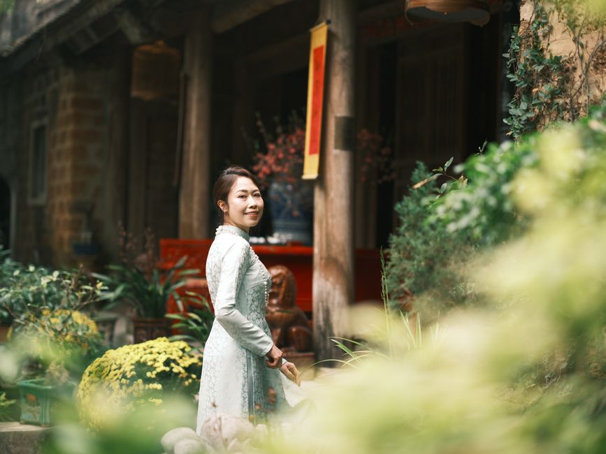 A woman in a traditional áo dài poses gracefully amidst lush greenery and a historical Vietnamese setting.