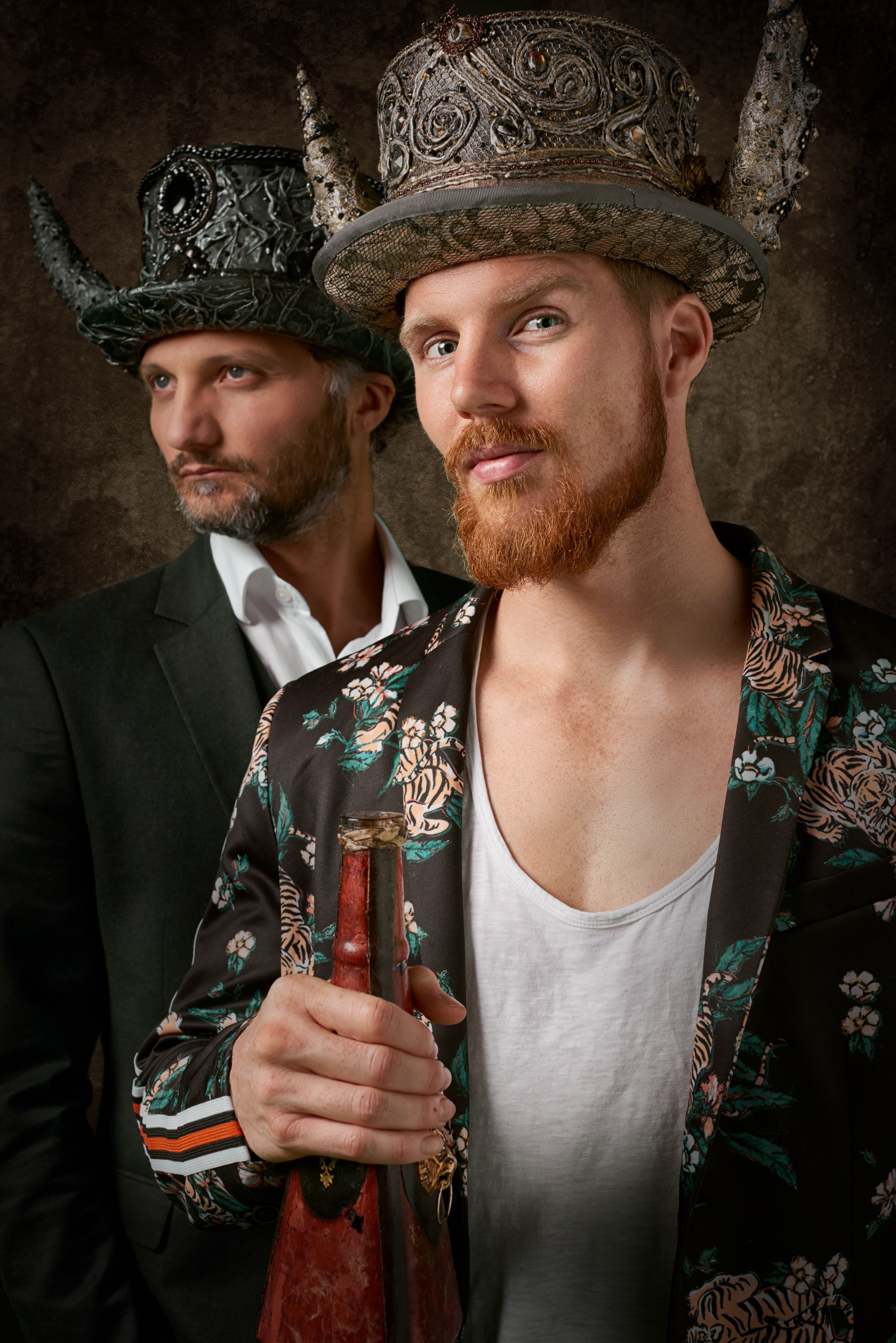 Two men in floral and ornate attire pose with decorative hats and bottles in a studio.
