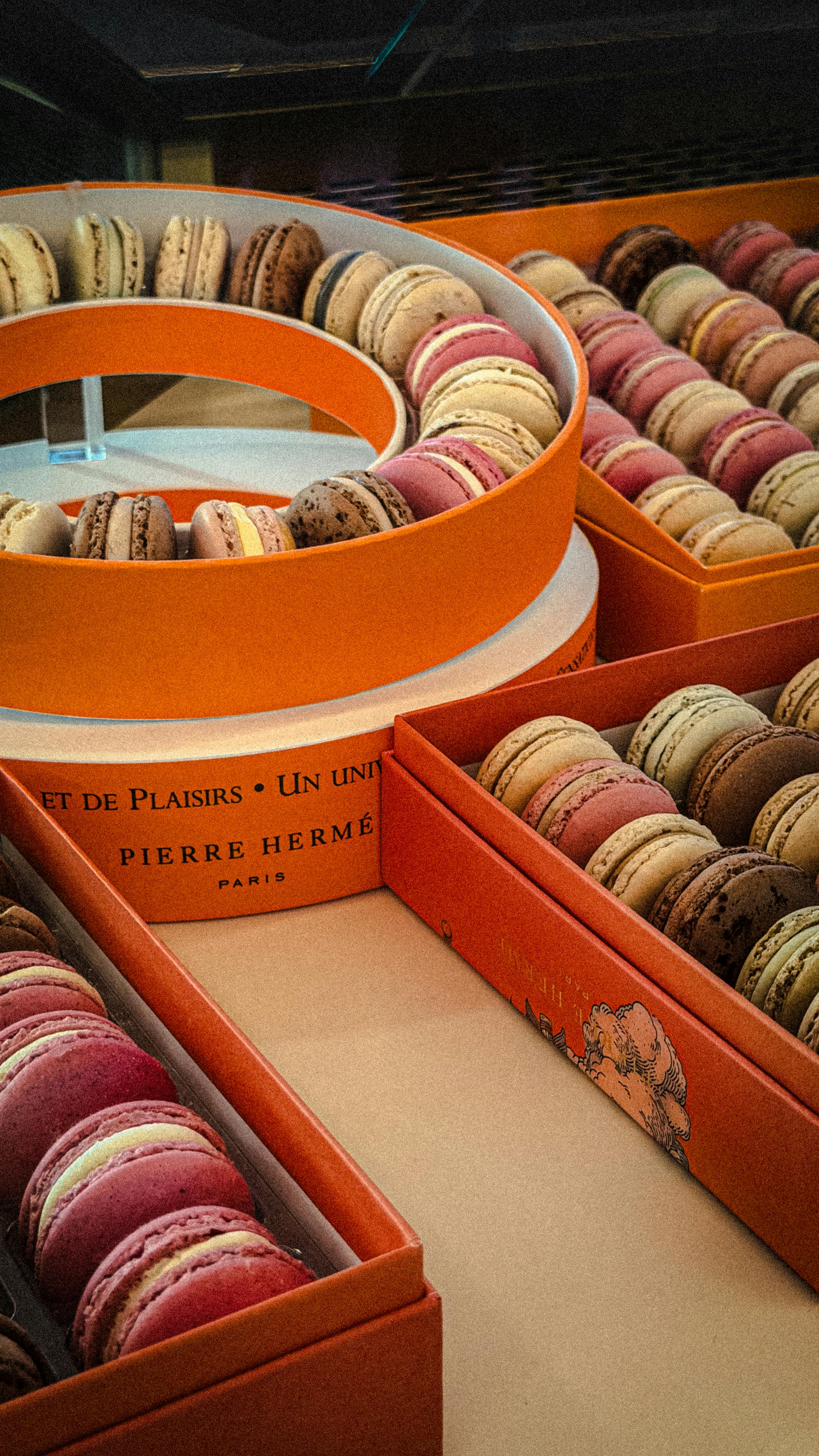 A vibrant display of assorted French macarons in elegant orange boxes from Pierre Hermé.
