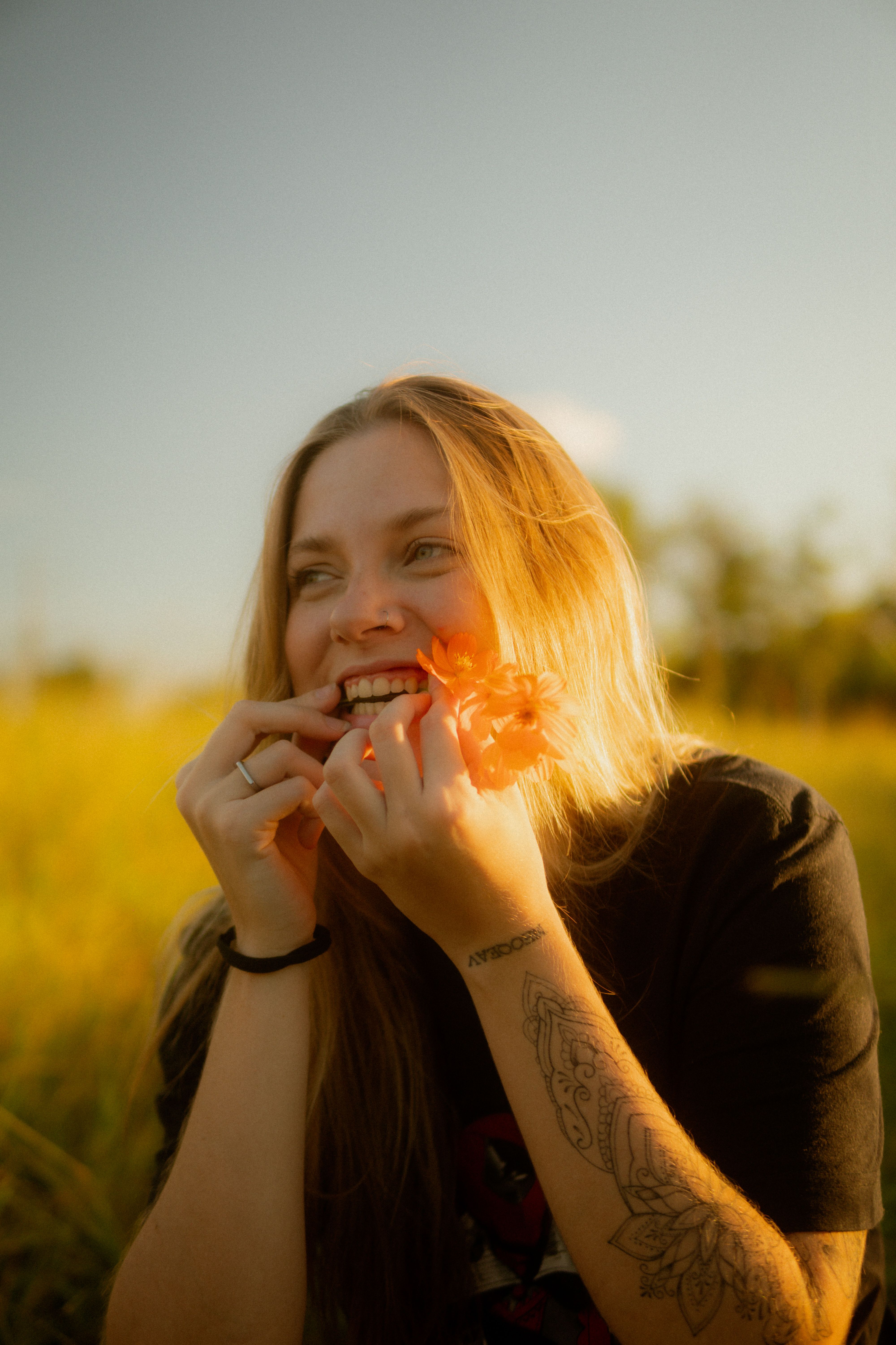 A young woman with tattoos smiles holding flowers in a sunlit field in Sete Lagoas, Brazil.