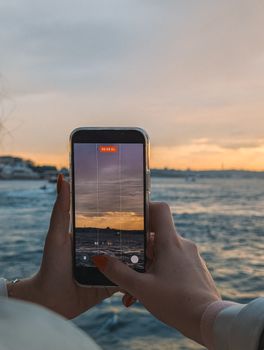 A close-up of hands holding a smartphone capturing a stunning İstanbul sunset over the sea.