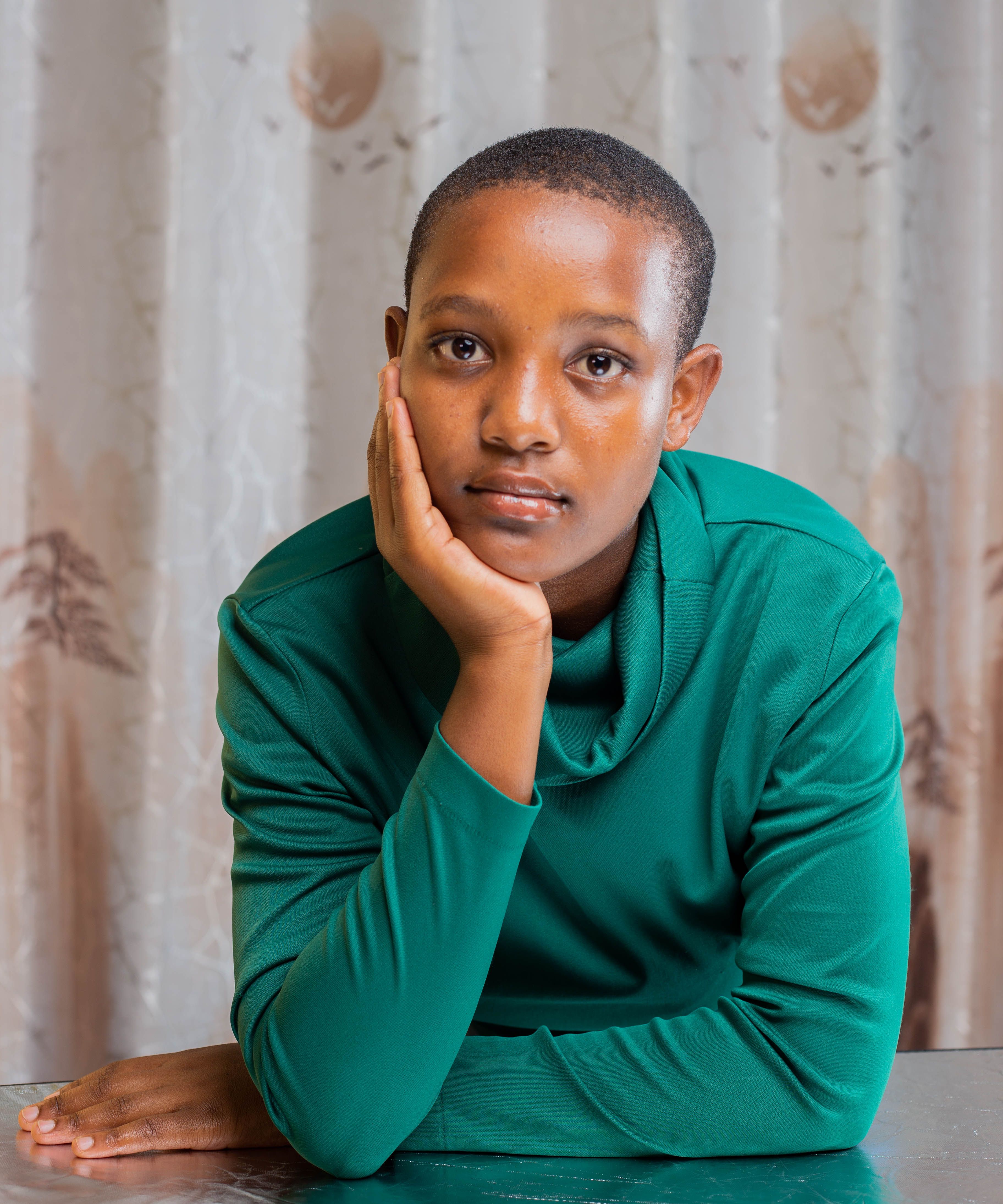 A young woman in a green top, resting her face on her hand, gazes thoughtfully indoors.