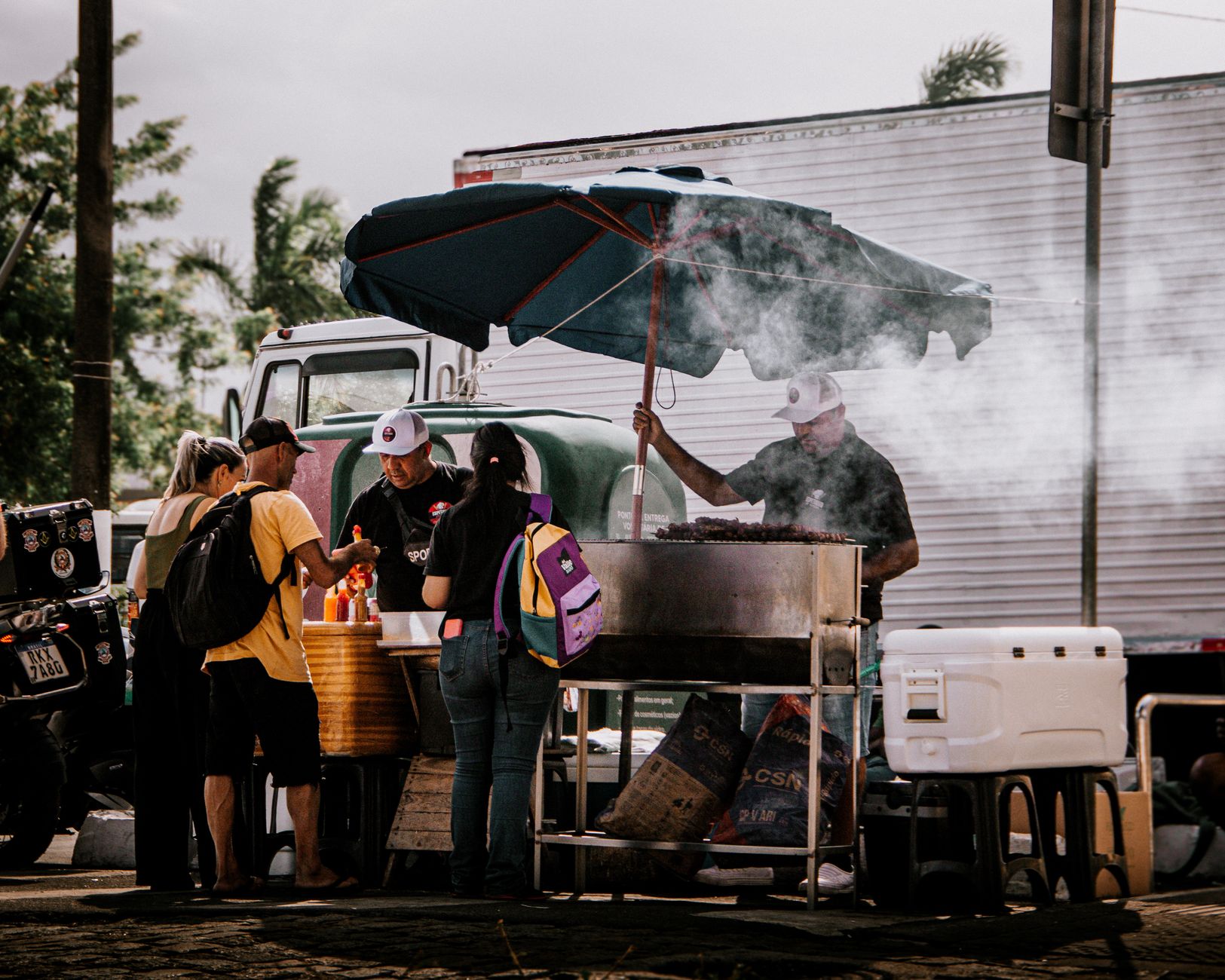 Street food vendor in action under umbrella serving customers. Smoky atmosphere adds charm.