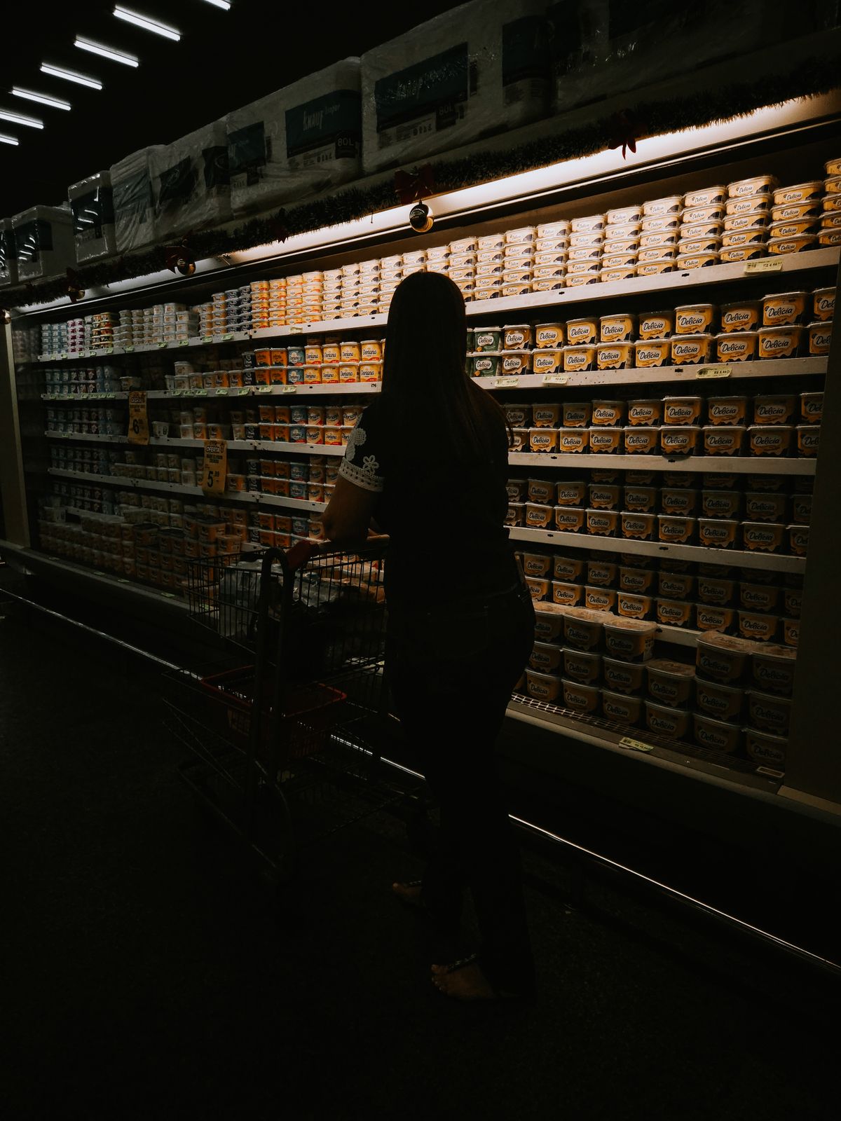 A woman with a shopping cart silhouetted against a lit dairy shelf in a dim supermarket aisle