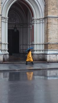 A person holding an umbrella walks by a Gothic-style building in rainy Novi Sad, Serbia.