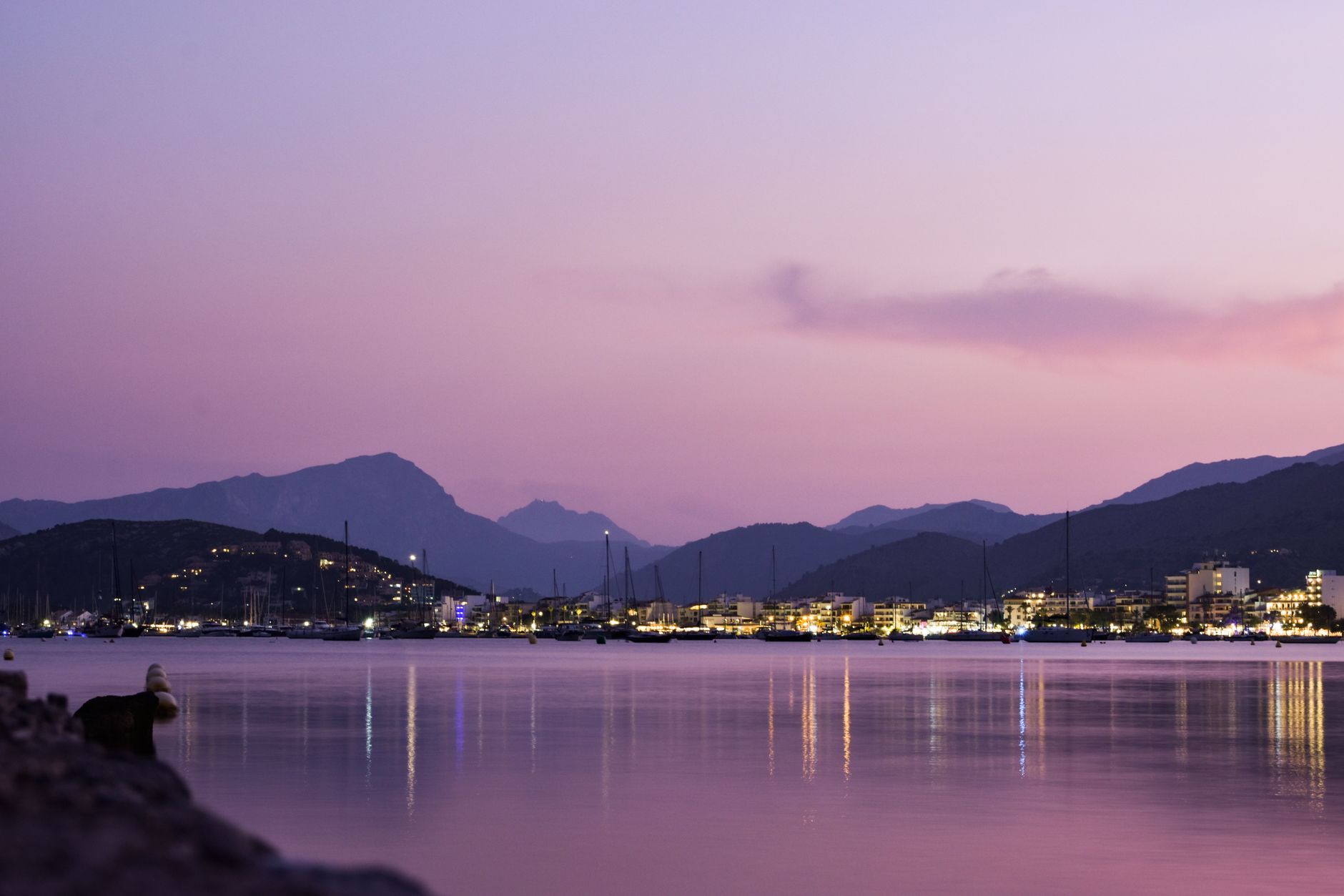 Serene twilight harbor scene with distant mountains and city lights reflecting on the calm water.