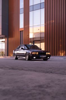 A sleek classic BMW E34 parked in front of a modern building at sunset, showcasing elegant design.