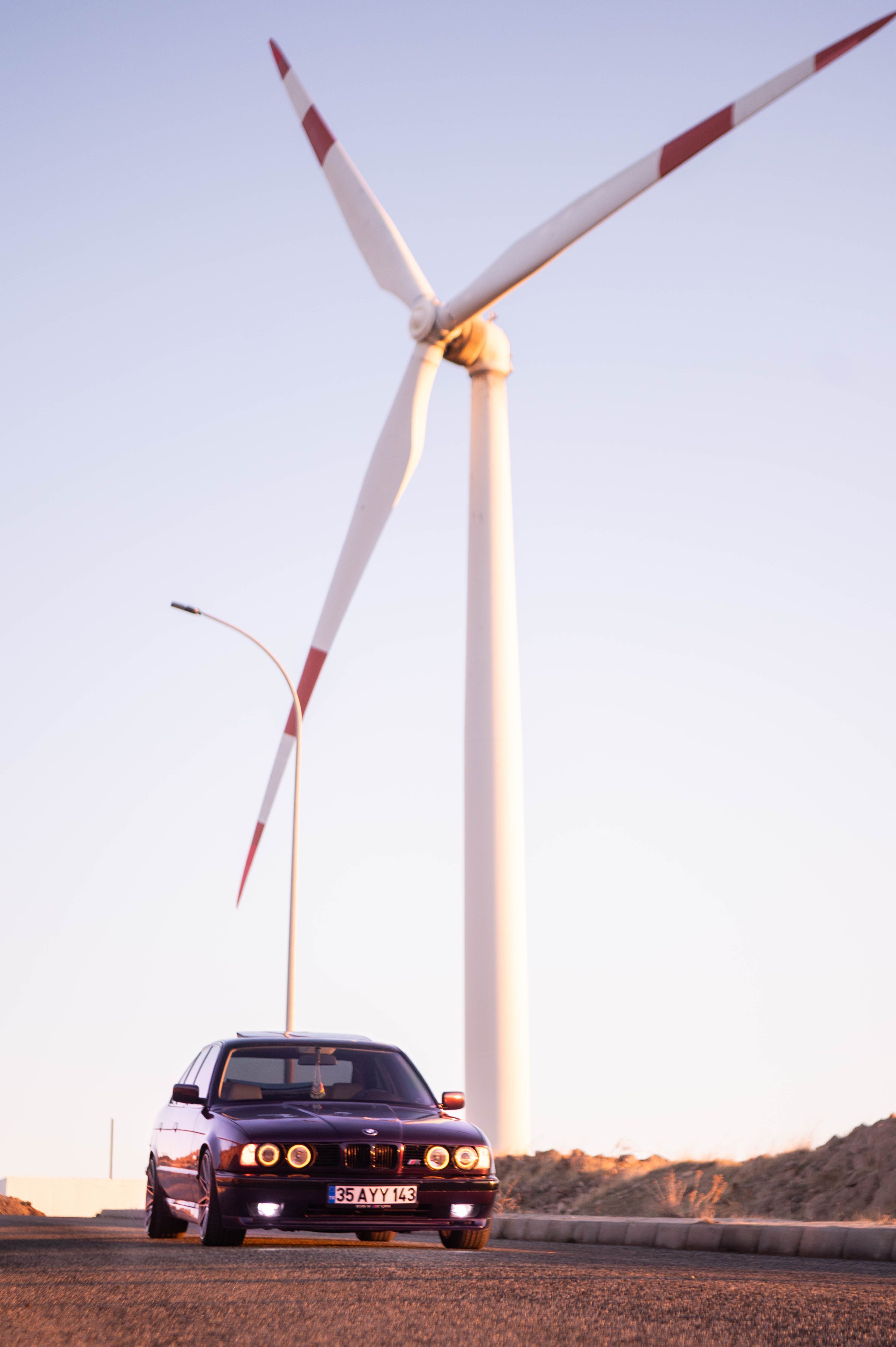 Classic BMW E34 driving near a towering wind turbine at sunset.