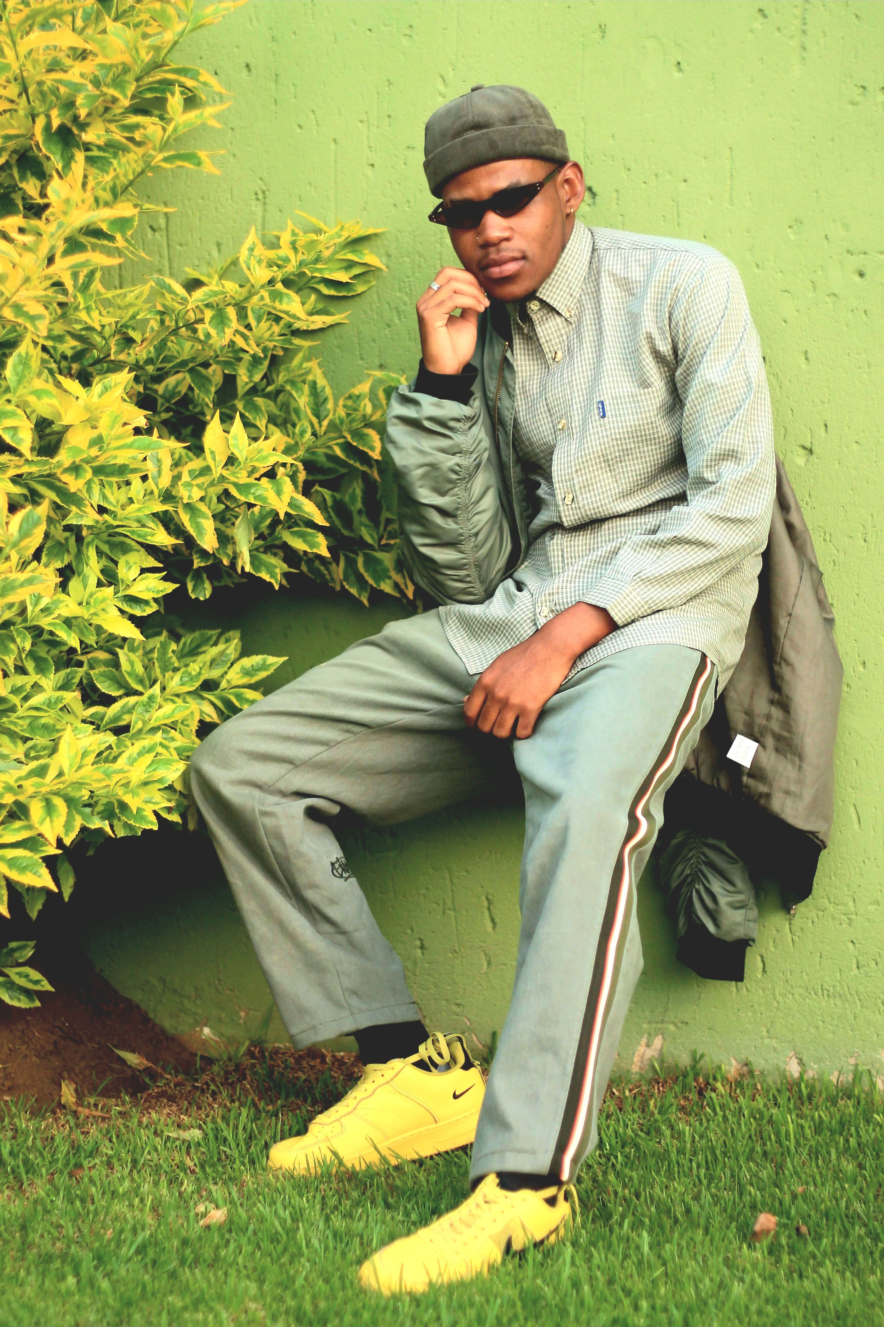 Fashionable man in sunglasses and casual attire posing near a green wall outdoors.