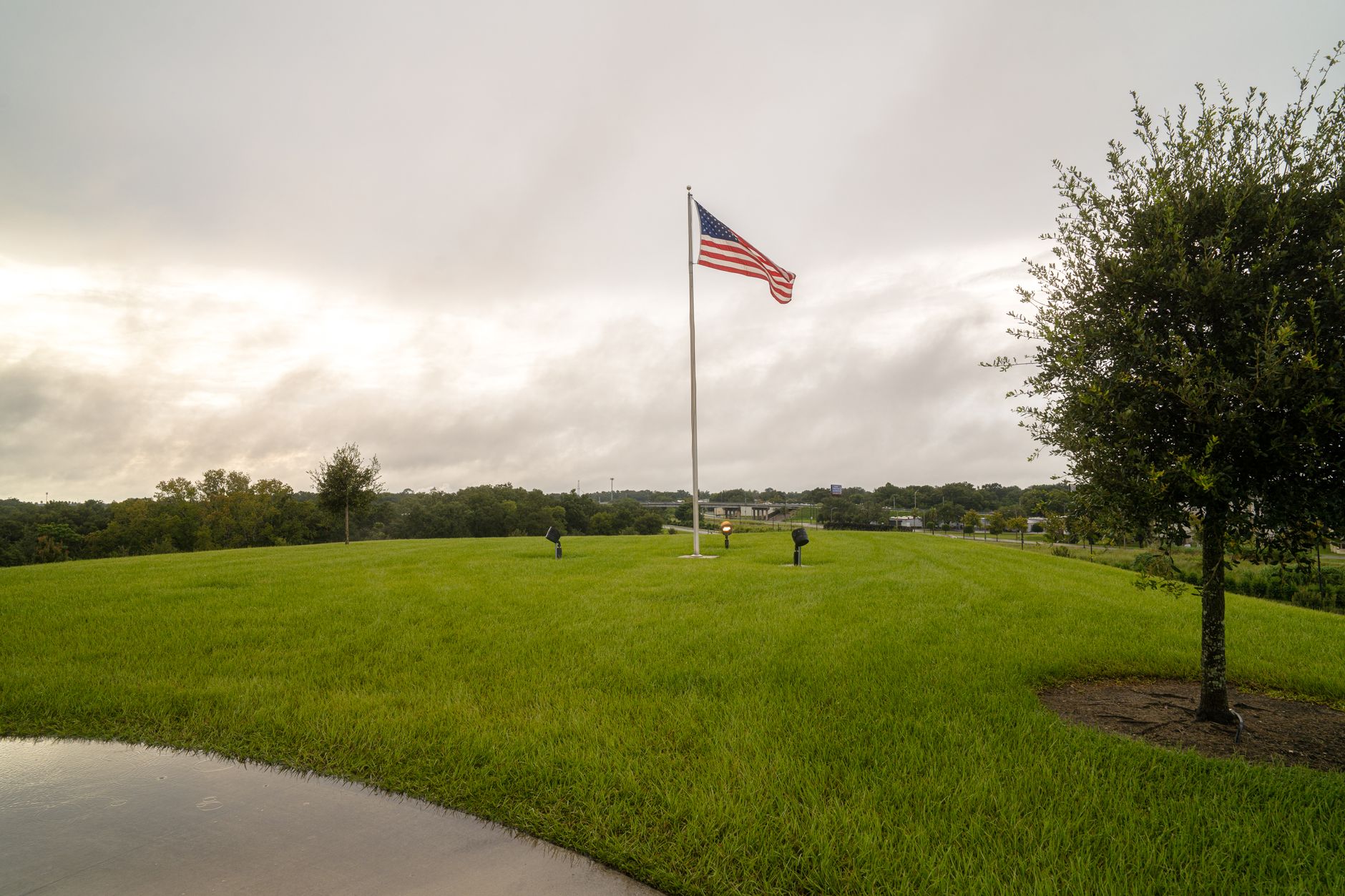 A prominent American flag stands tall on a lush green lawn beneath a cloudy, overcast sky.