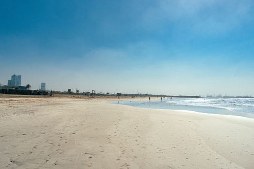 Free Expansive beach view in Sharjah, UAE with a vibrant blue sky and few people enjoying the shoreline. Stock Photo