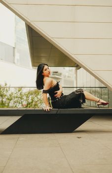 Elegant woman in black dress poses on a sleek bench in a contemporary urban setting, showcasing style.