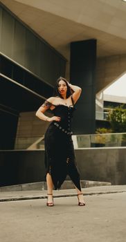 A stylish fashion model striking a pose in a modern urban environment, wearing a black dress.