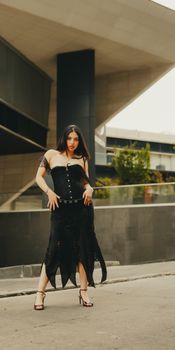 Stylish woman in a black dress and heels posing in an urban setting, showcasing modern fashion.