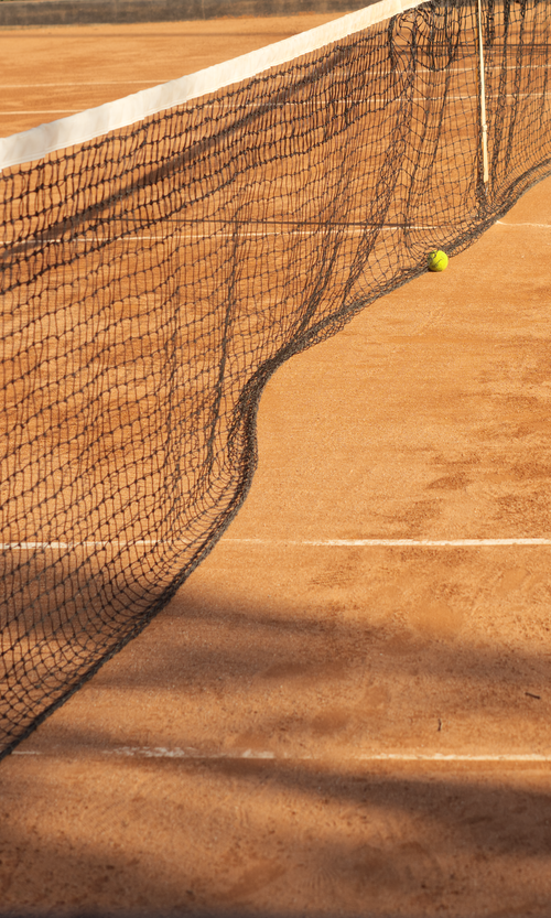Free A close-up of a tennis ball resting on a clay court next to the net in bright sunlight. Stock Photo
