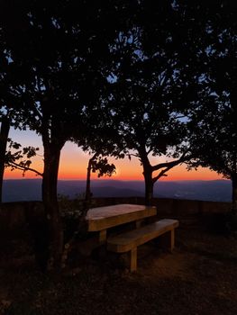 Silhouetted trees and benches against a vibrant sunset sky create a serene outdoor scene.