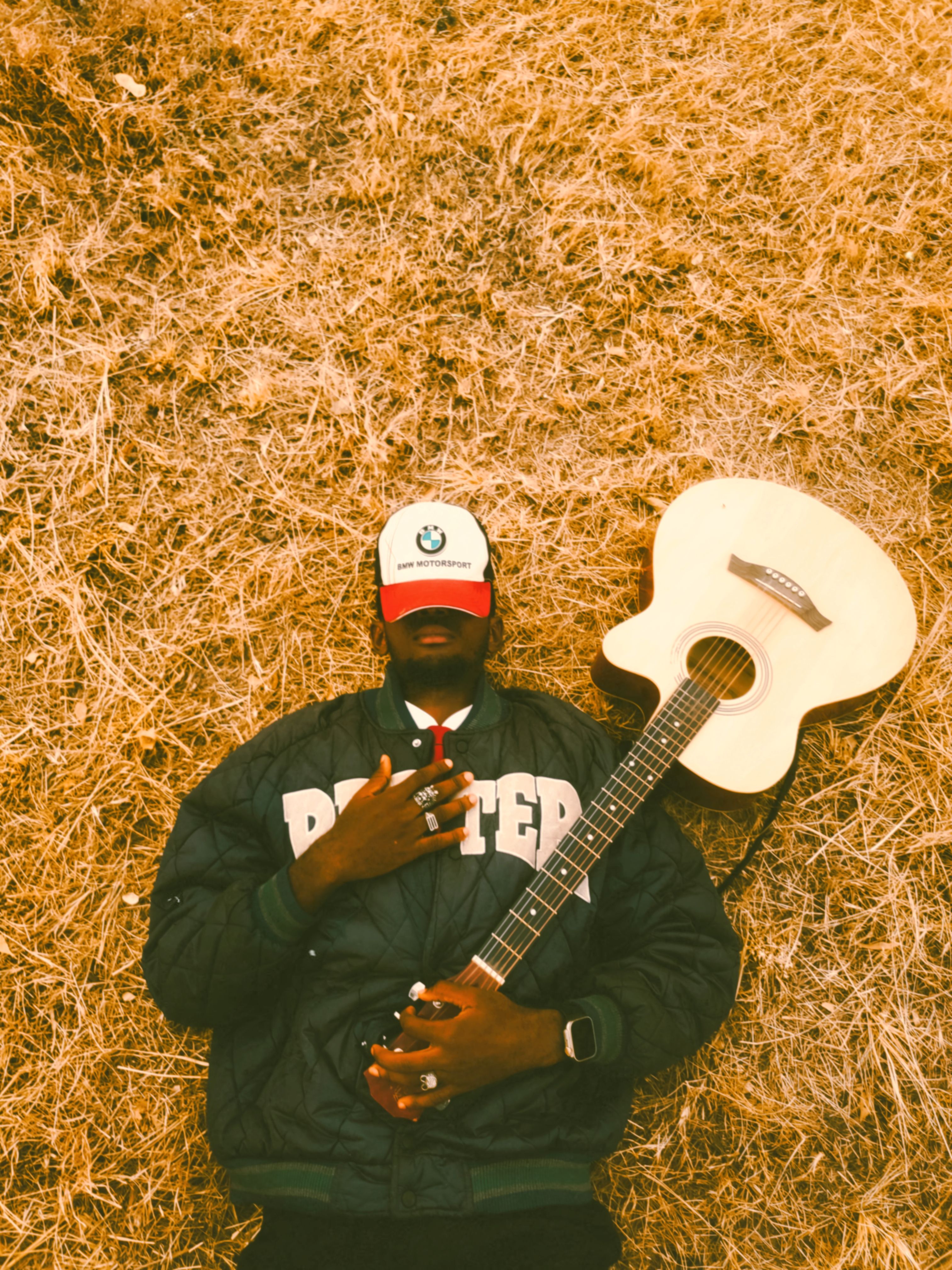 A person in a jacket and cap lying on dry grass with an acoustic guitar.