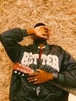 Man in quilted jacket lying on dry grass with guitar, outdoors in autumn setting