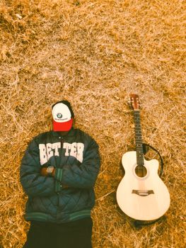 Man in casual wear lying on dry grass with an acoustic guitar beside him.
