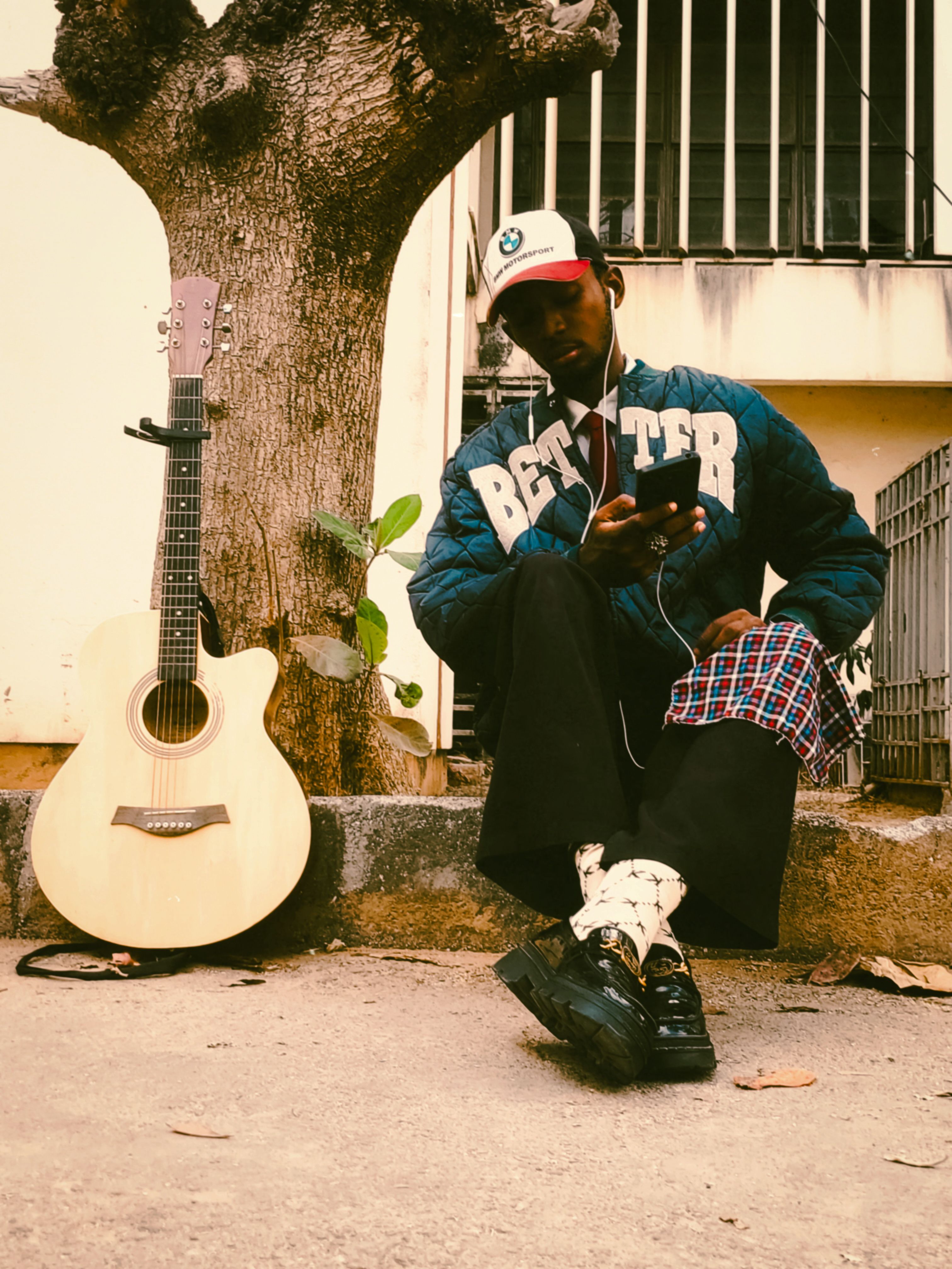 A young man leaning against a tree, listening to music with a guitar nearby.