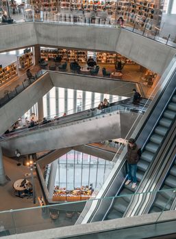 Interior view of a modern multi-level library with escalators and shelves filled with books, bustling with people.