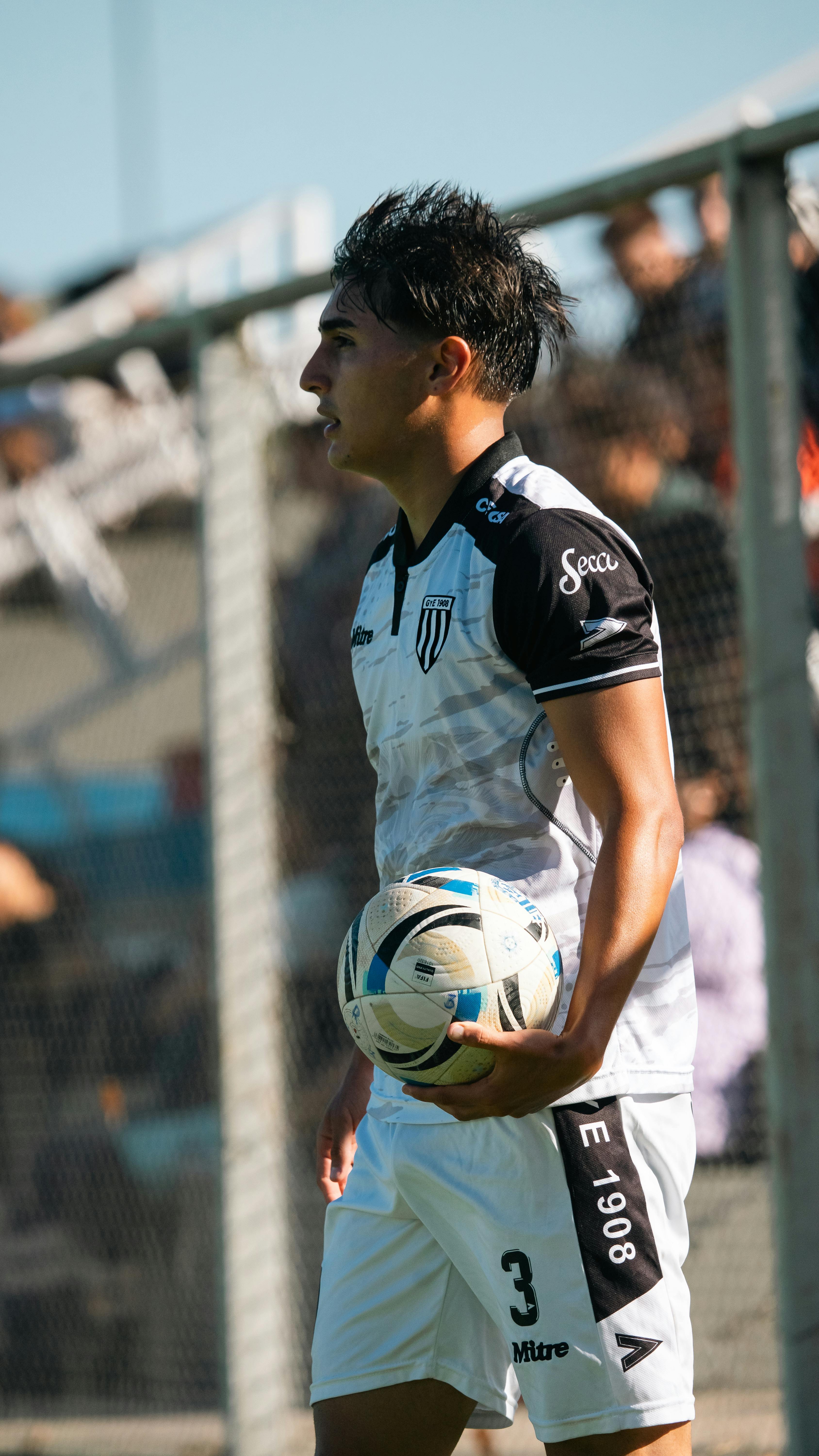 A soccer player in a black and white uniform holds a ball on a sunny day, ready to throw in.