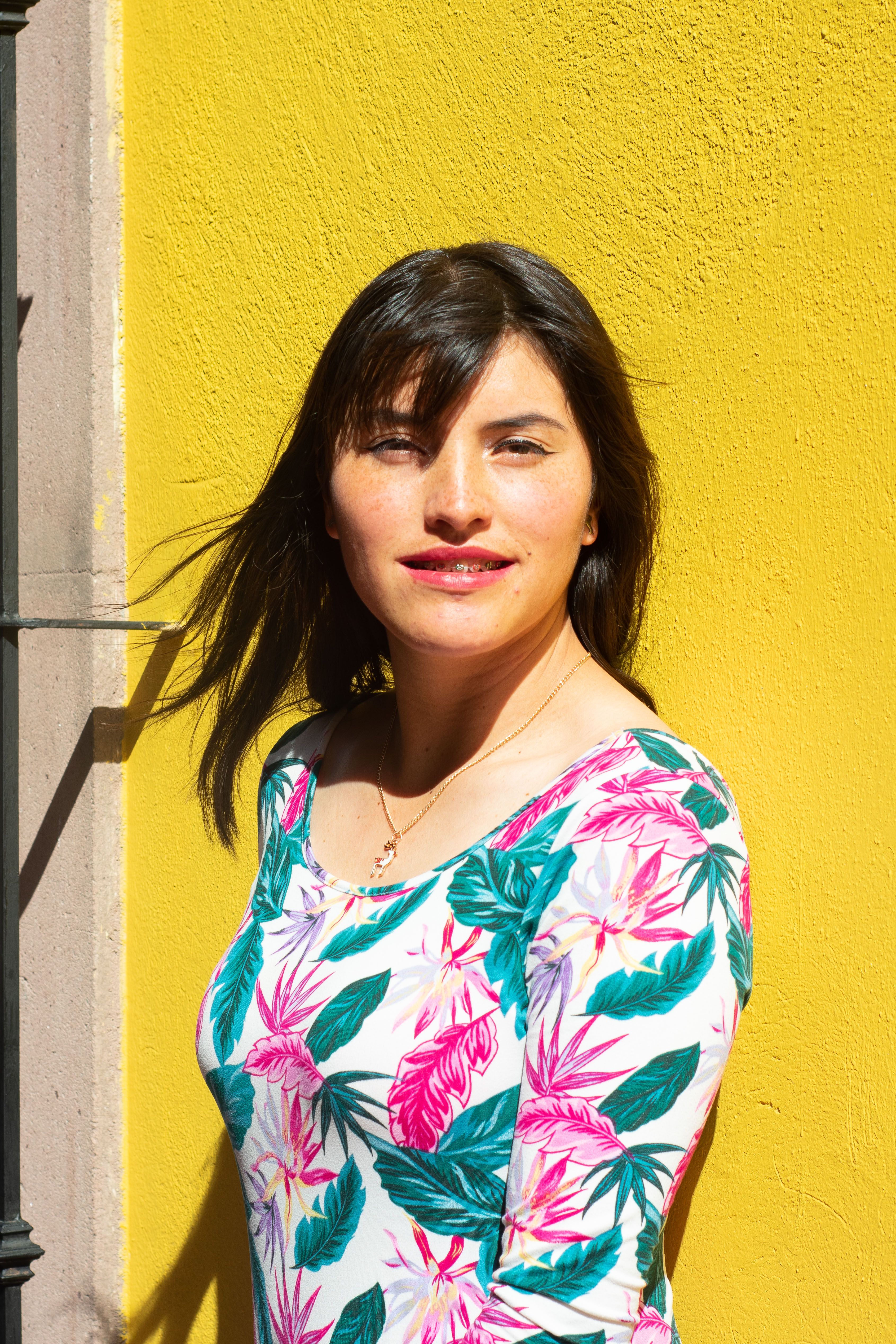 A woman stands against a vibrant yellow wall in Lerma, Mexico, wearing a floral dress, captured in natural light.