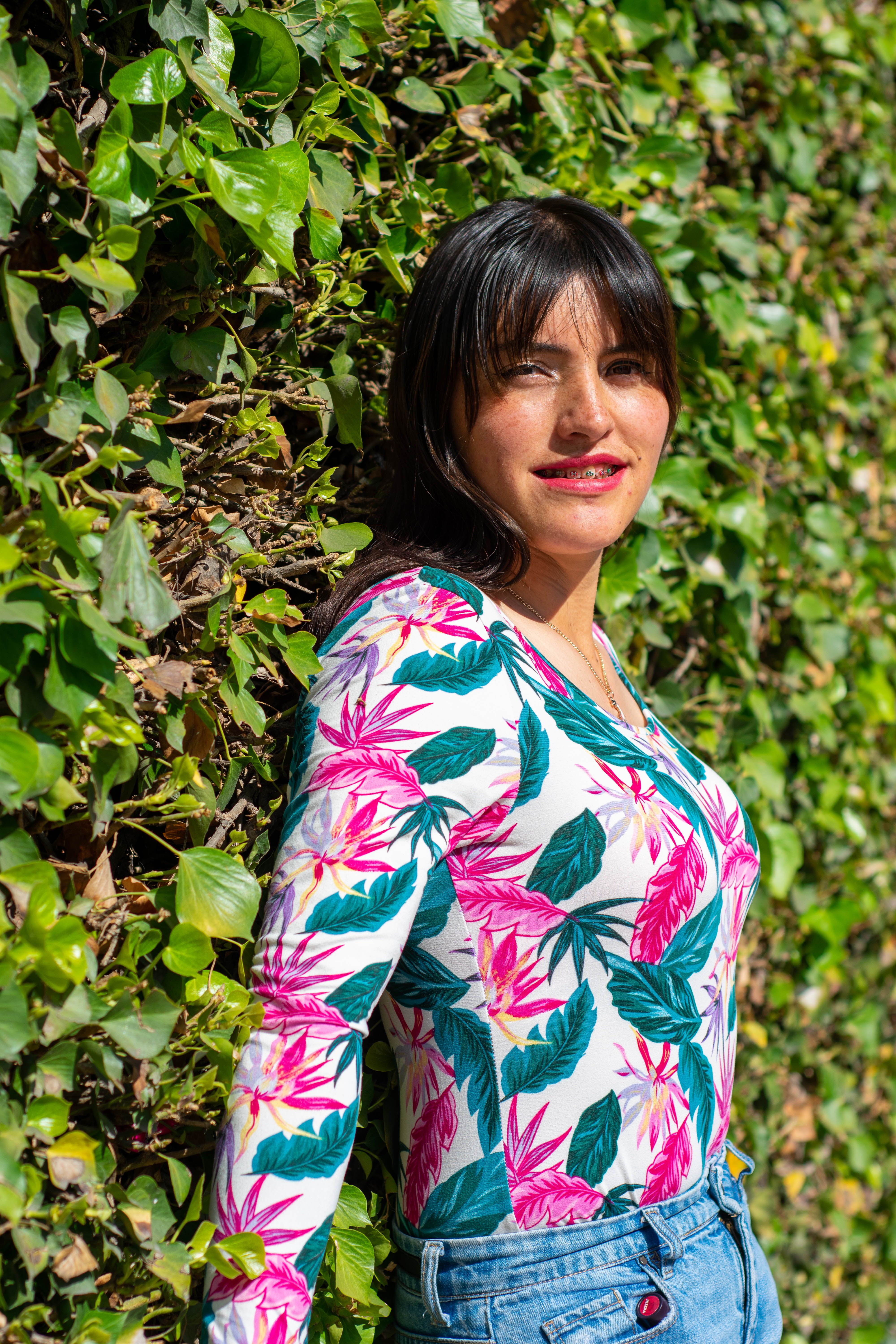 Portrait of a woman wearing a floral top against an ivy-covered wall in sunlight.