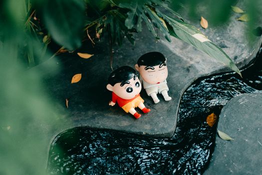 Two cartoon figurines sit on a stone ledge near a lush garden stream.
