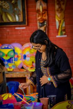 A woman artist creatively paints vibrant masks in a studio filled with colorful artwork.