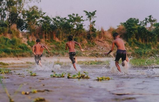 Three boys running in a river, splashing water during sunset, full of joy and energy.