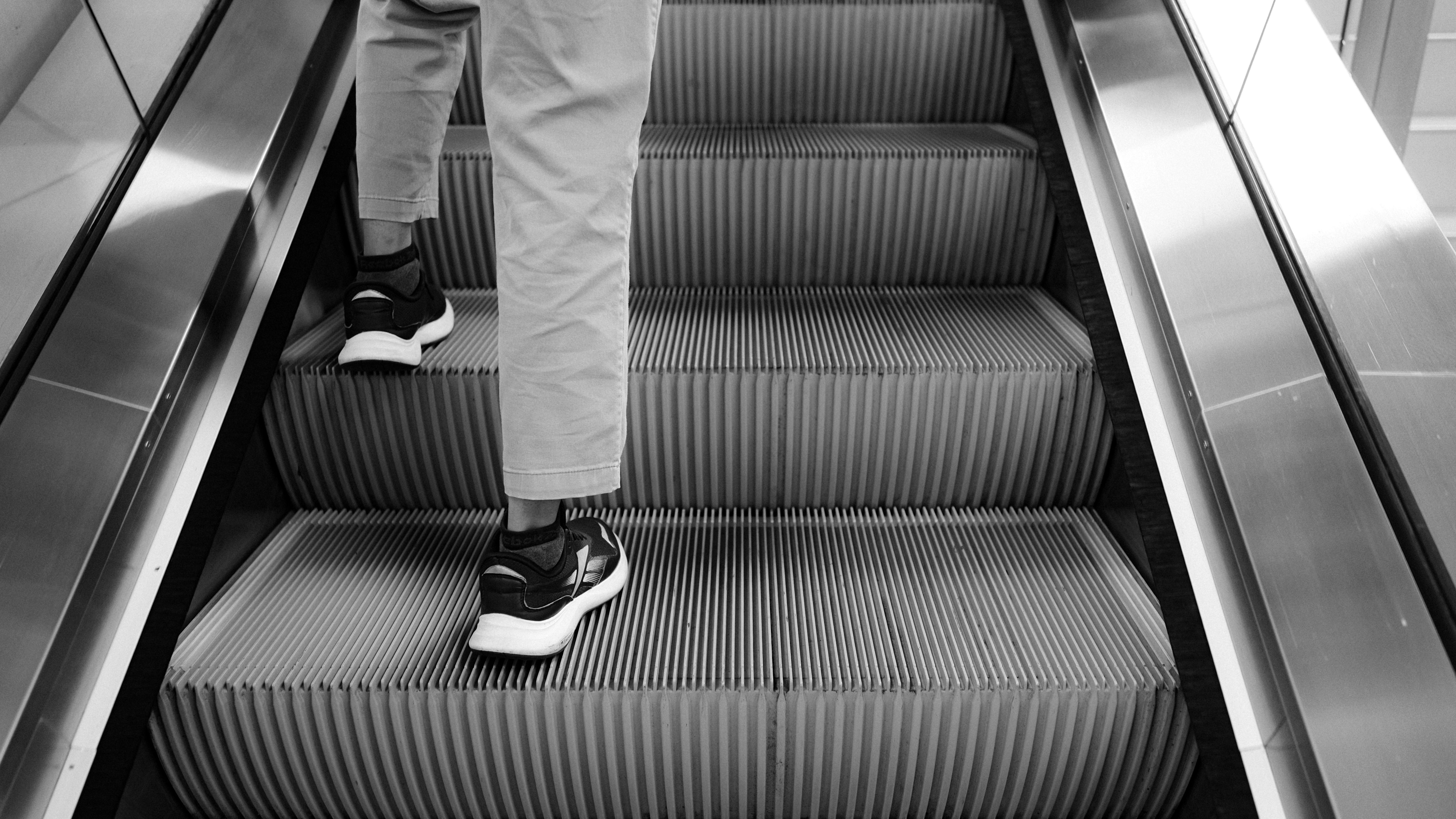 白黒の背景の中で、スニーカーを履いた人がエスカレーターの上に立っている。