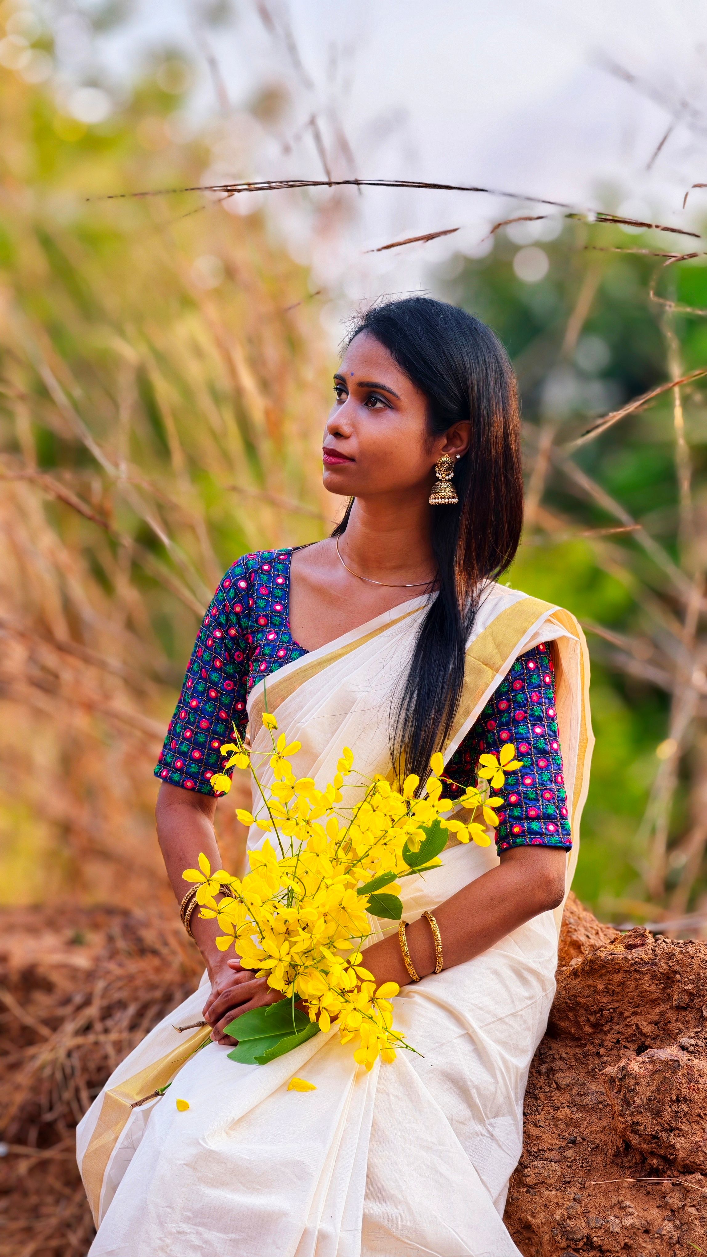 A serene portrait of a South Asian woman in a traditional Kasavu saree holding yellow flowers outdoors.