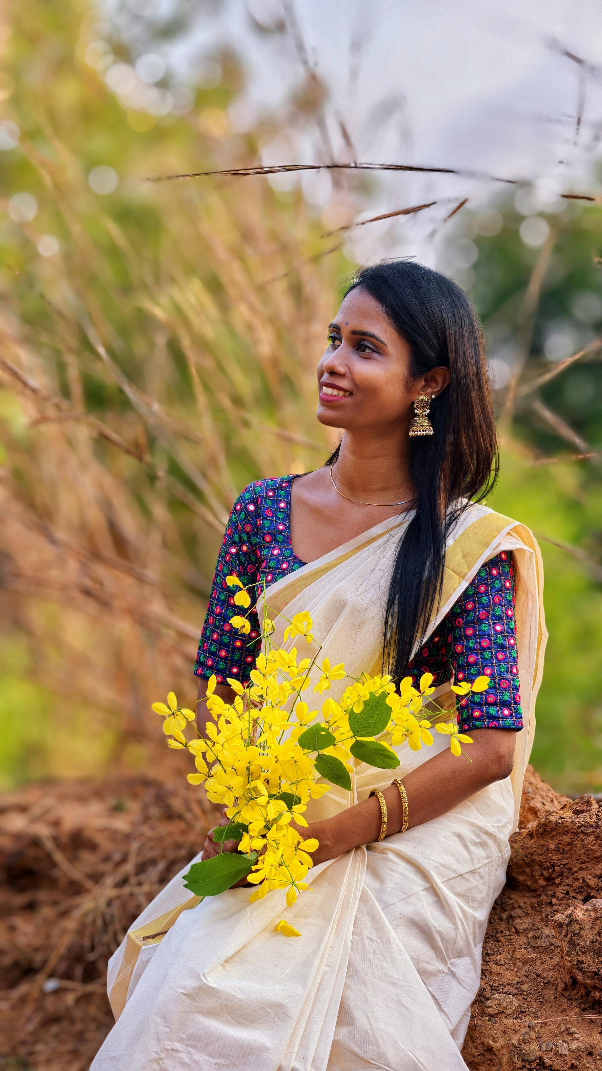 A South Asian woman in traditional attire holding yellow flowers in a serene outdoor setting.