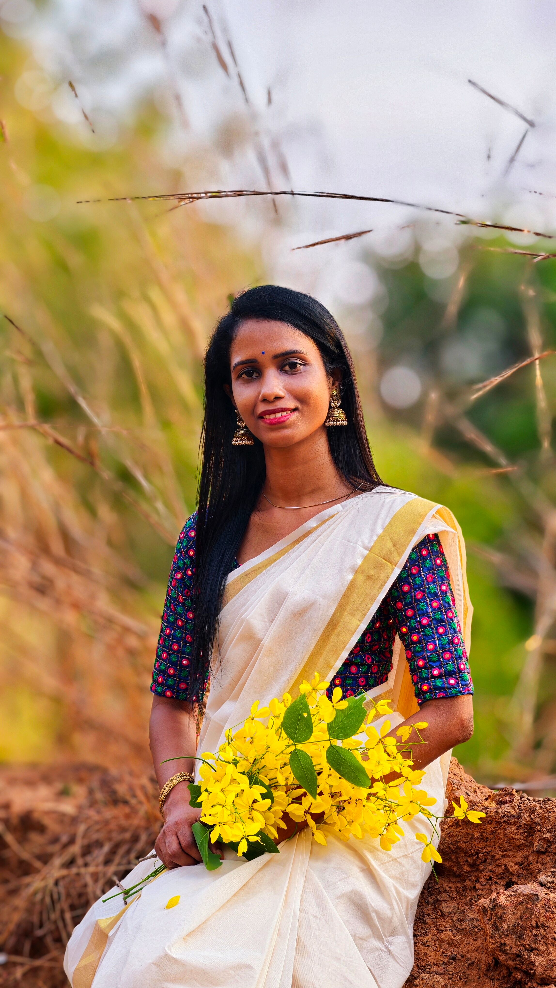 A South Indian woman in traditional Kerala attire holding Kanikkonna flowers outdoors.