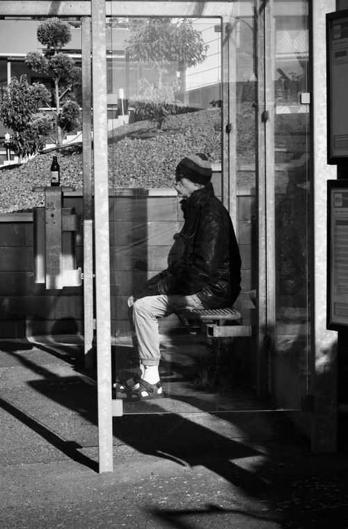 Free Black and white photo of an elderly man patiently sitting at a bus stop, capturing a moment of solitude. Stock Photo