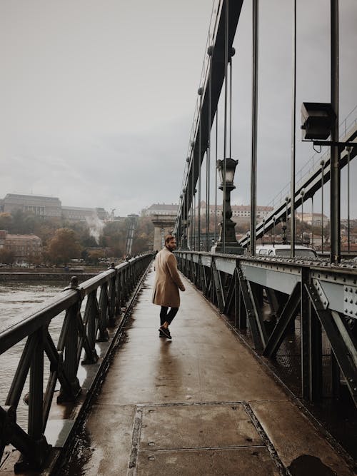 Man in Brown Coat and Black Pants Walking on Bridge