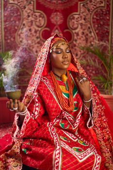 Serene Nigerian woman in vibrant traditional attire meditates indoors, with incense smoke creating a mystical ambiance.