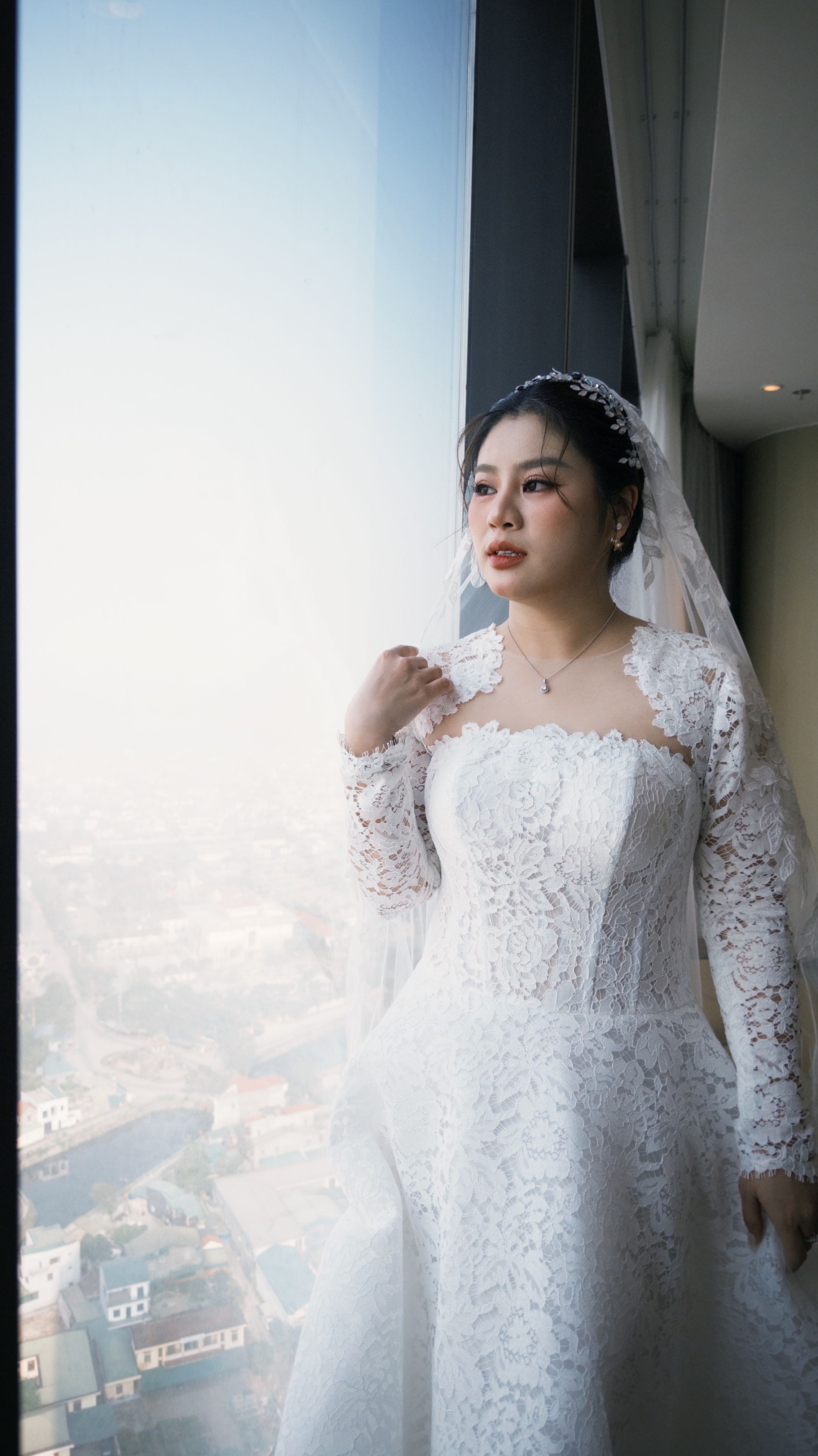 Vietnamese bride gazing out a window in a luxurious lace gown, portraying elegance and tradition.