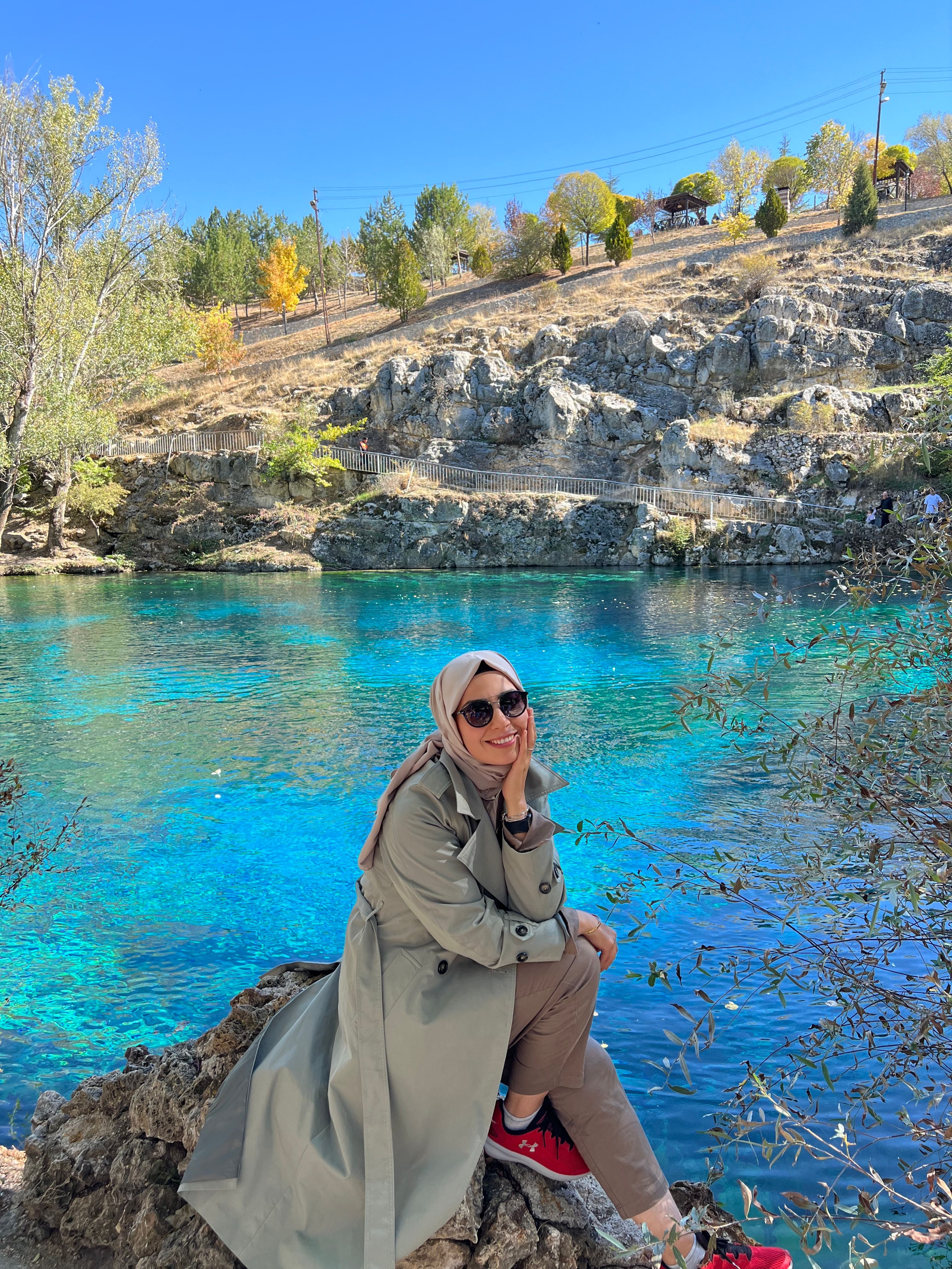 Relaxed woman in hijab by vibrant blue lake amidst autumn foliage.