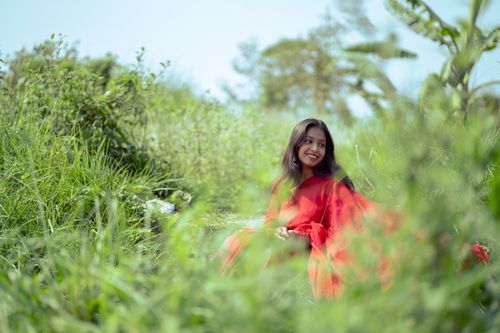 Free A woman in a vibrant red saree sitting joyfully amidst lush greenery in a serene outdoor setting. Stock Photo