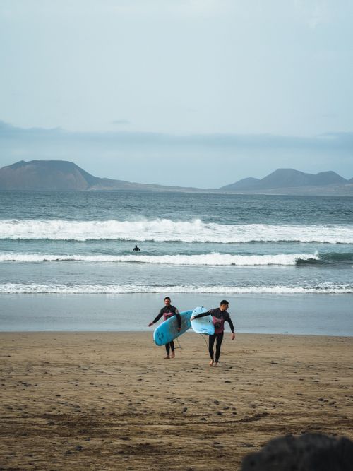 Free Surfers walking along the sandy beach in Lanzarote, Spain, carrying surfboards against ocean waves backdrop. Stock Photo