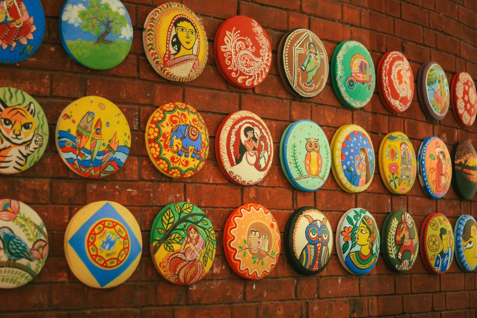 Vibrant hand-painted plates with cultural motifs displayed on a brick wall in Dhaka.