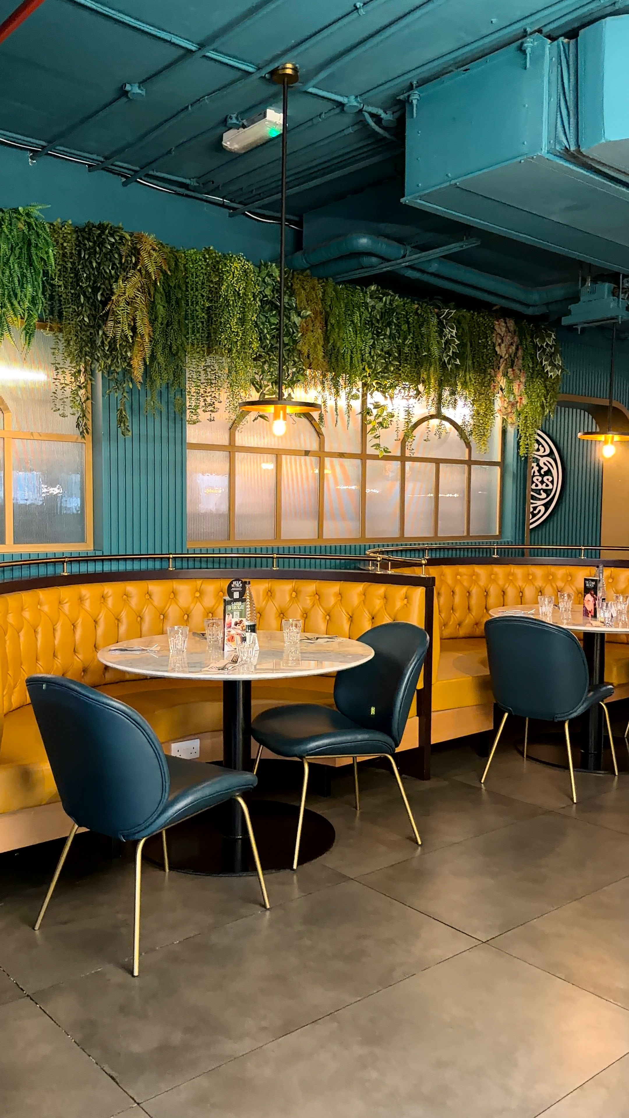 Chic restaurant interior featuring yellow booths and blue chairs, under hanging greenery.