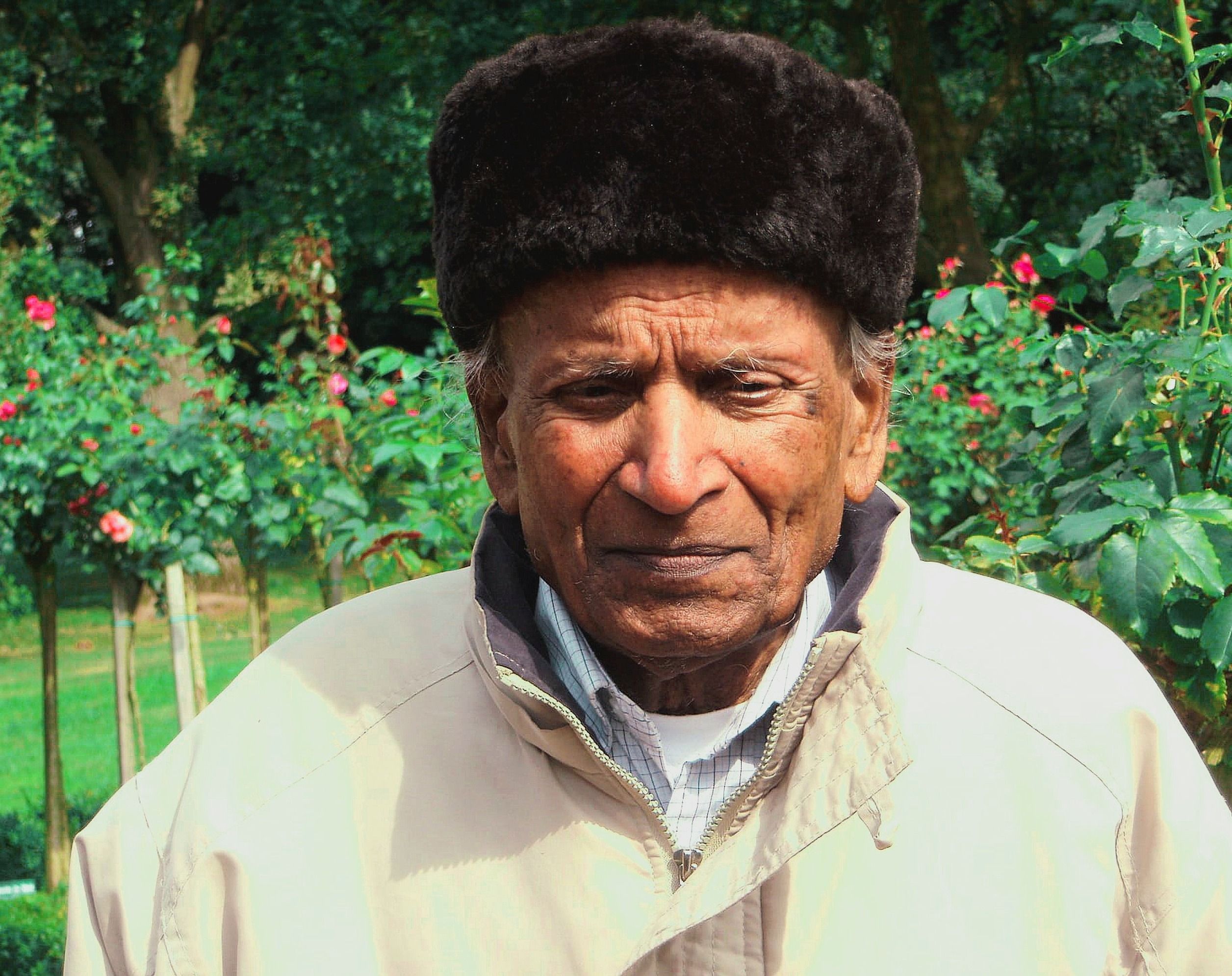 Senior man wearing a warm hat in a lush garden setting, surrounded by flowers.