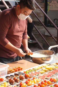 A street vendor prepares colorful skewered desserts under daylight, showcasing vibrant fruits.