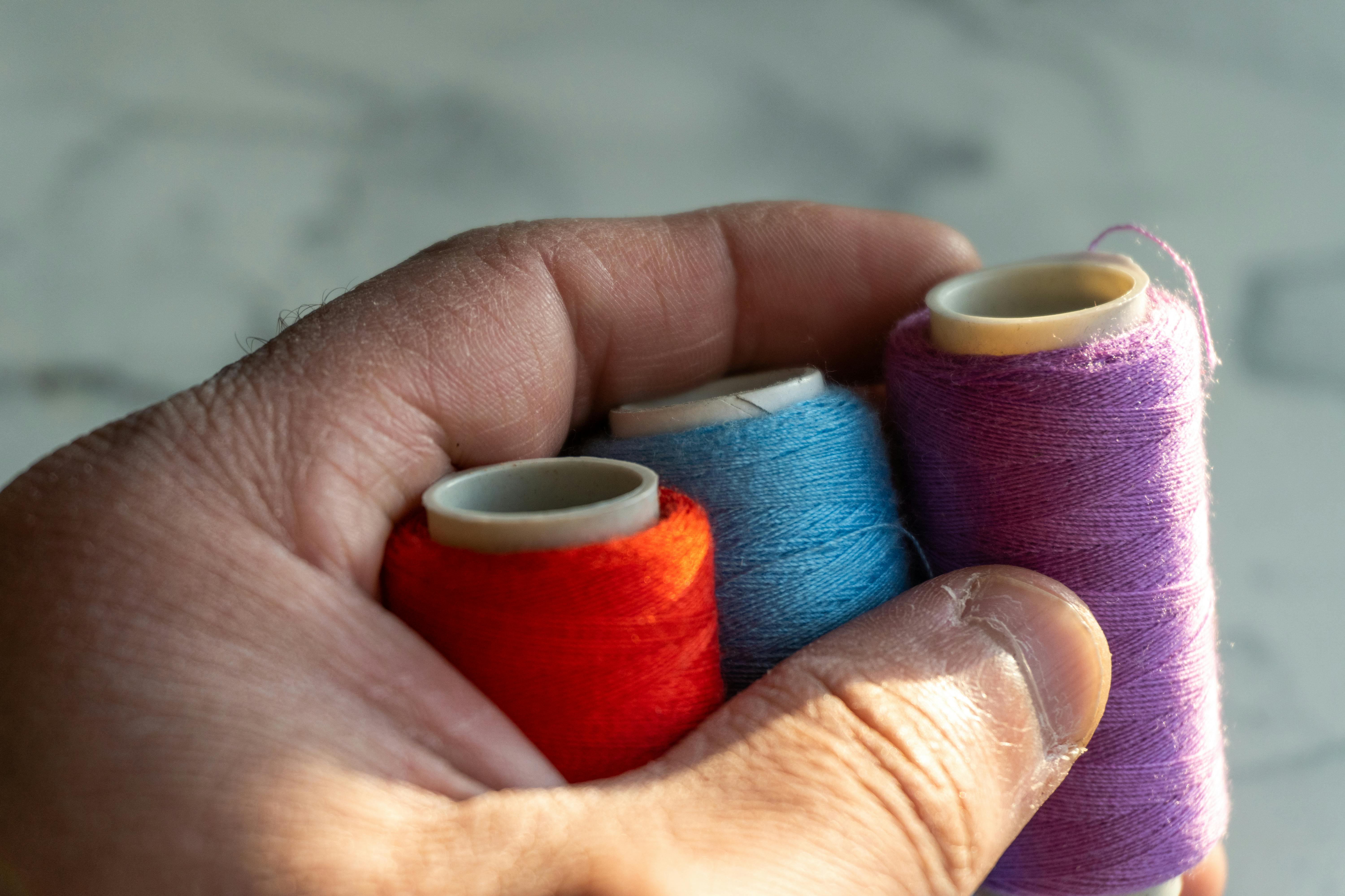 Close-up of human hand holding red, blue, and purple sewing threads with natural lighting. — Maynard Longarm Quilting