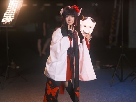 A cosplayer in traditional costume holding an oni mask, set against a dimly lit indoor scene.
