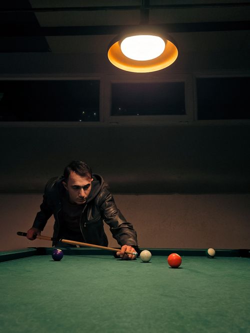 Free A focused man lines up a shot on a pool table under a warm light in a dim interior setting. Stock Photo
