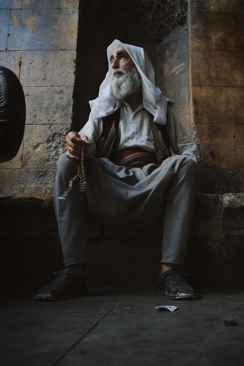 Free An elderly man in traditional attire sits in a dimly lit alleyway, creating a serene and reflective atmosphere. Stock Photo